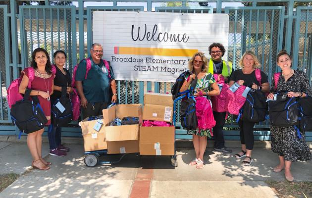 people holding backbacks, standing next to boxes of backpacks for donation