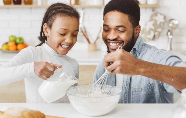 A photo of a smiling father and daughter pouring milk into a mixing bowl.