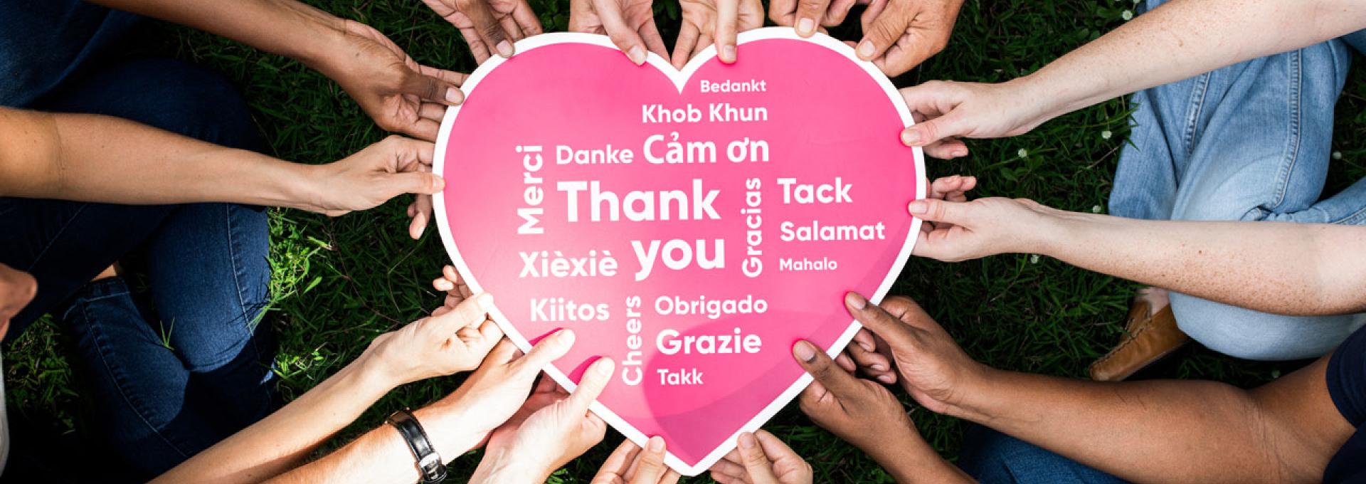 Group of people holding a heart-shaped sign that displays "Thank you" in different languages.